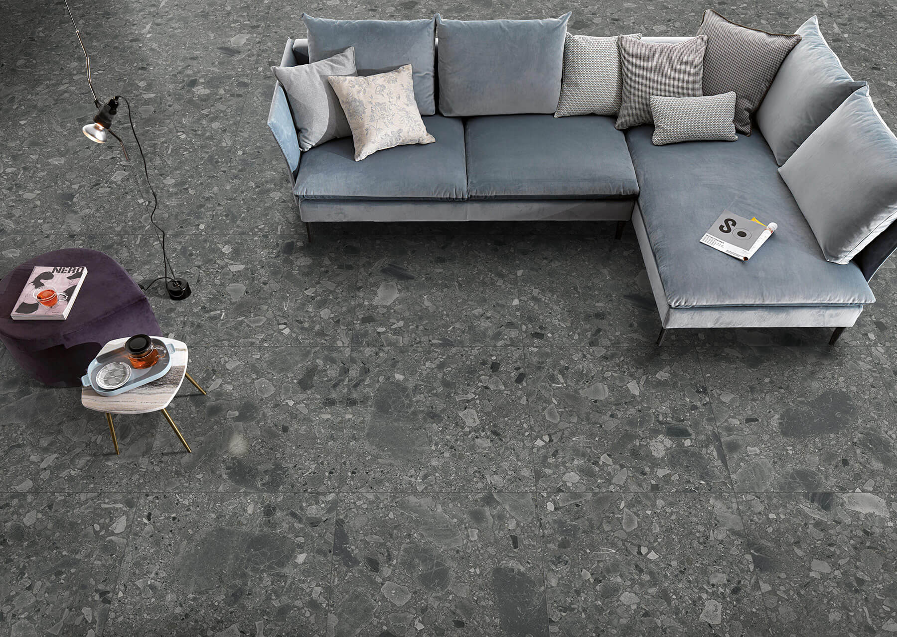 Looking down on a display lounge area with silver velvet corner couch, small purple velvet coffee table and feature flooring of large charcoal grey tiles in a concrete stone style.