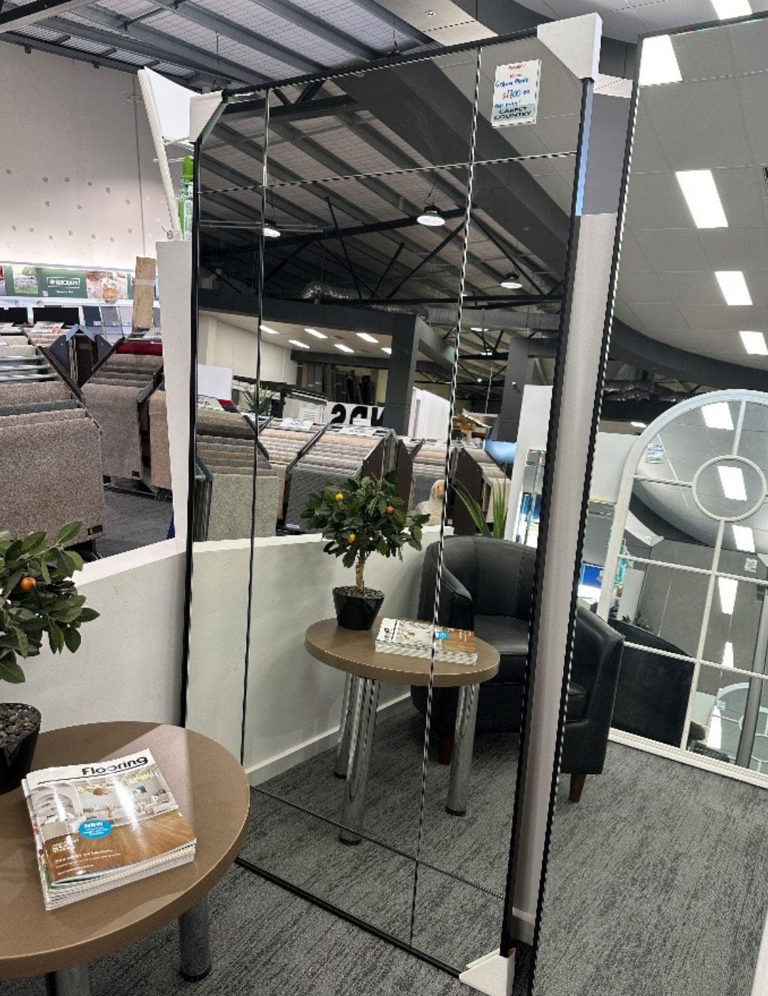 Tall rectangle mirror with black edge on display in a store. The mirror is reflecting other parts of the store like carpet on display and other mirrors.