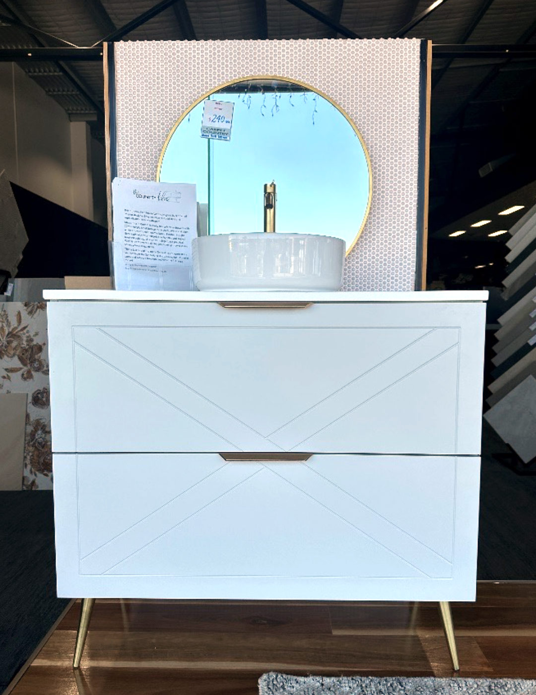 While farmhouse vanity with two drawers, and a white sink, gold tap and gold edged round mirror all on display in a store window.