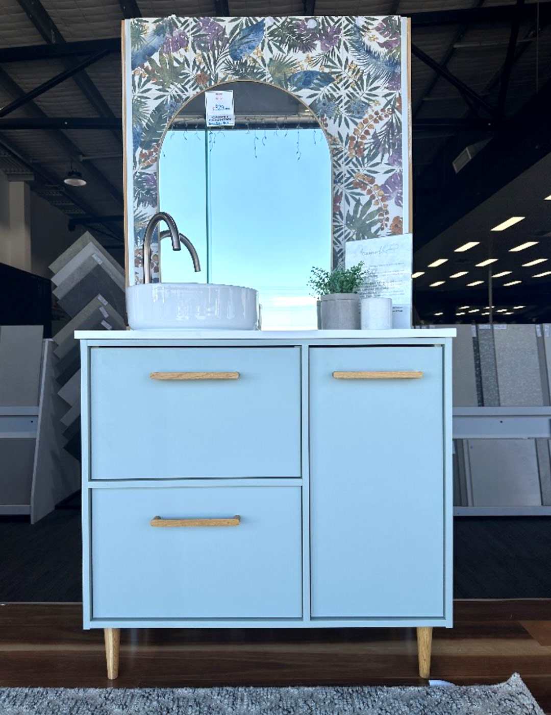 Light blue vanity in a store, it has three drawers, white top, wooden handles, and a white sink. A mirror with rounded top is behind it.