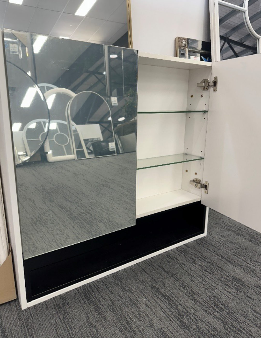 Mirror cabinet for a bathroom with two doors, the right door is open and shows three glass shelves in the cabinet. The cabinet is on the floor in a store.