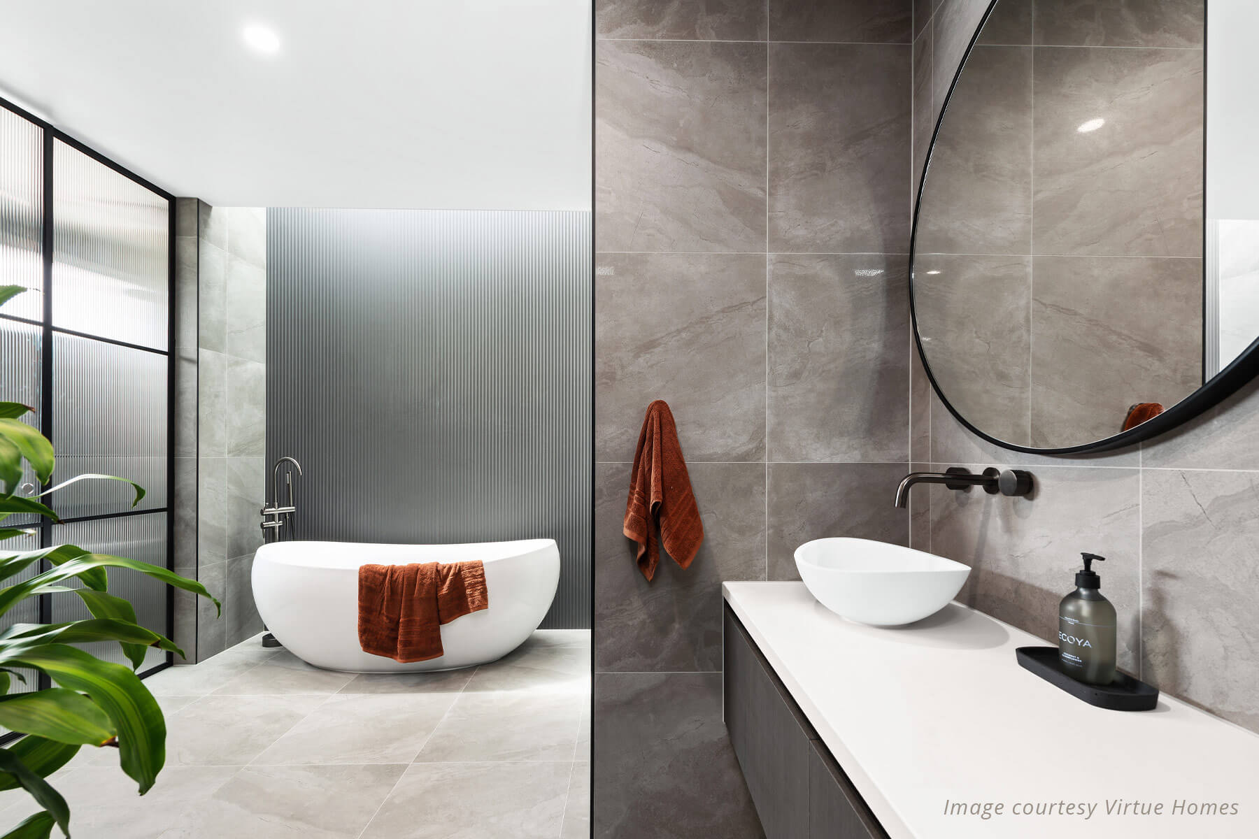 Luxury ensuite with white soaker tub and timber vanity. Grey tiles and large round mirror.