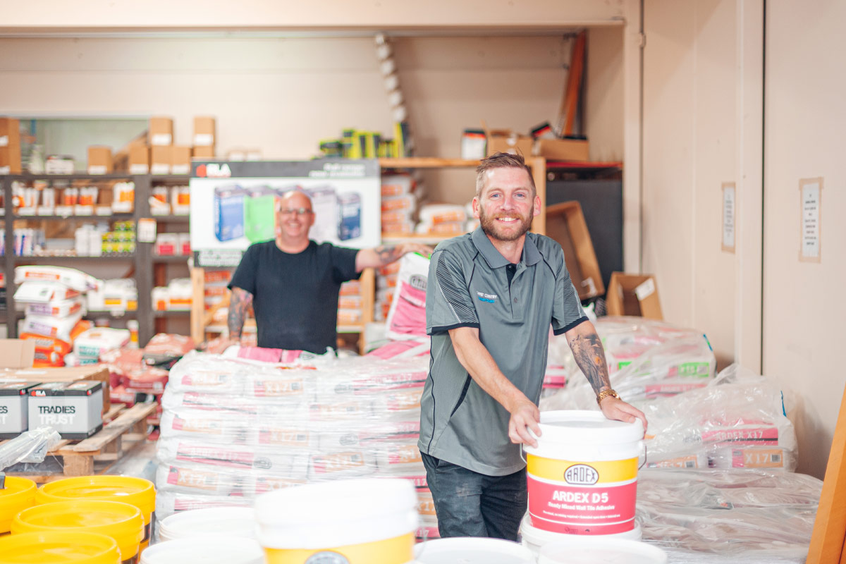 Two male staff in a tile shop surrounded by tile supplies like buckets of glue and grout.
