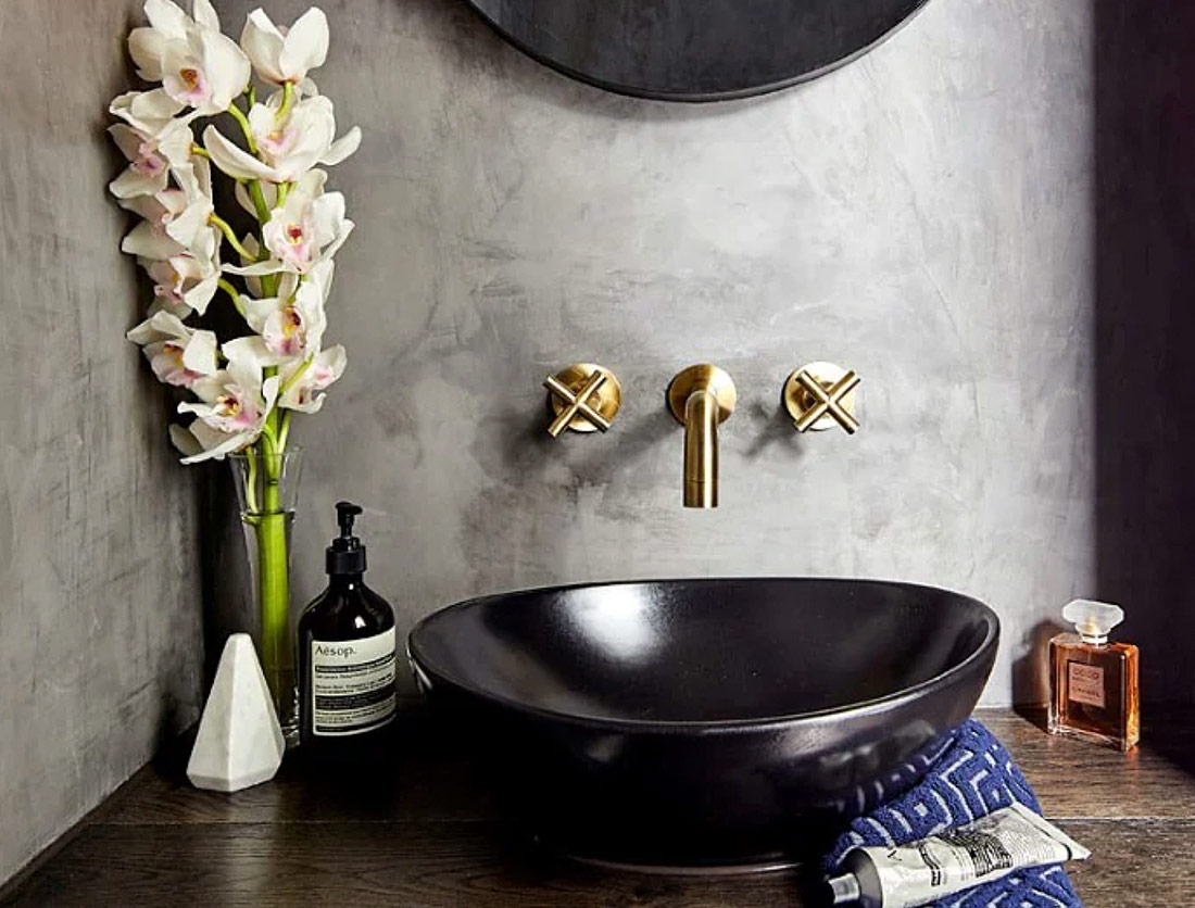 Close up of black basin and gold tapware, concrete walls in a bathroom.