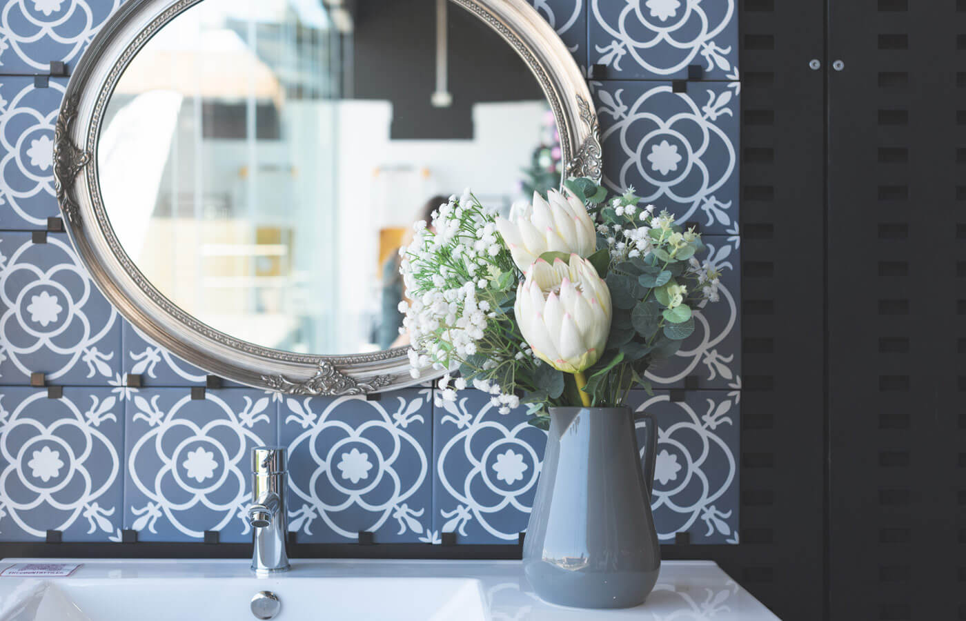 Large oval mirror with silver ornate frame hanging on display wall. Grey/blue and white floral tiles are behind the mirror, a white sink, silver tap, and grey vase with white flowers is on the vanity benchtop.
