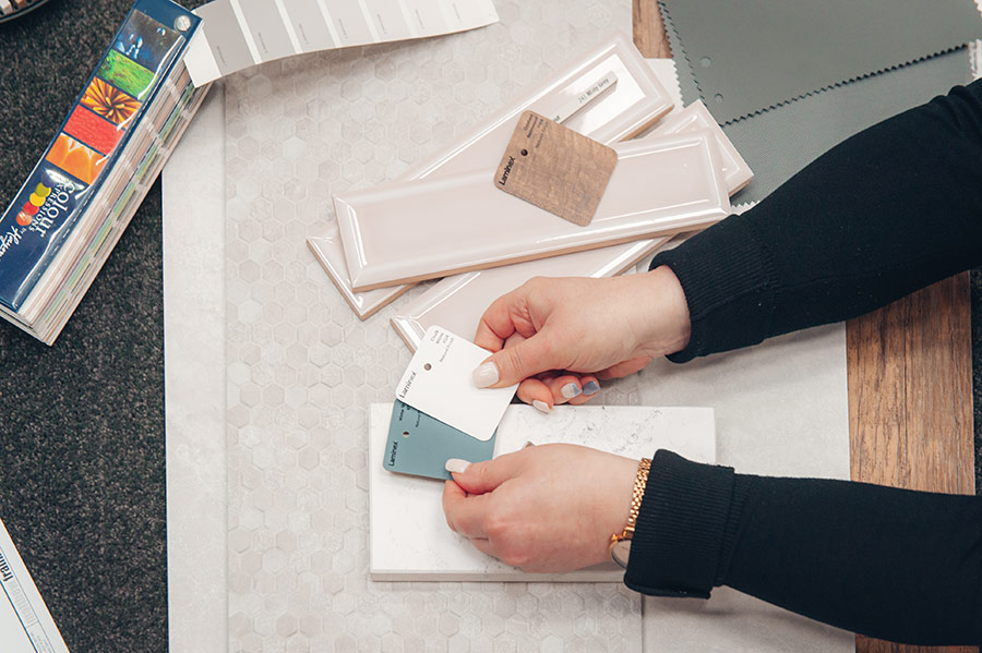 Woman wearing long sleeve black shirt and godl watch is holding a green and white colour swatch, comparing them to a number of tile samples.